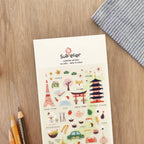 Sheet of stickers with various illustrations on a wooden surface with pencils and a cloth.